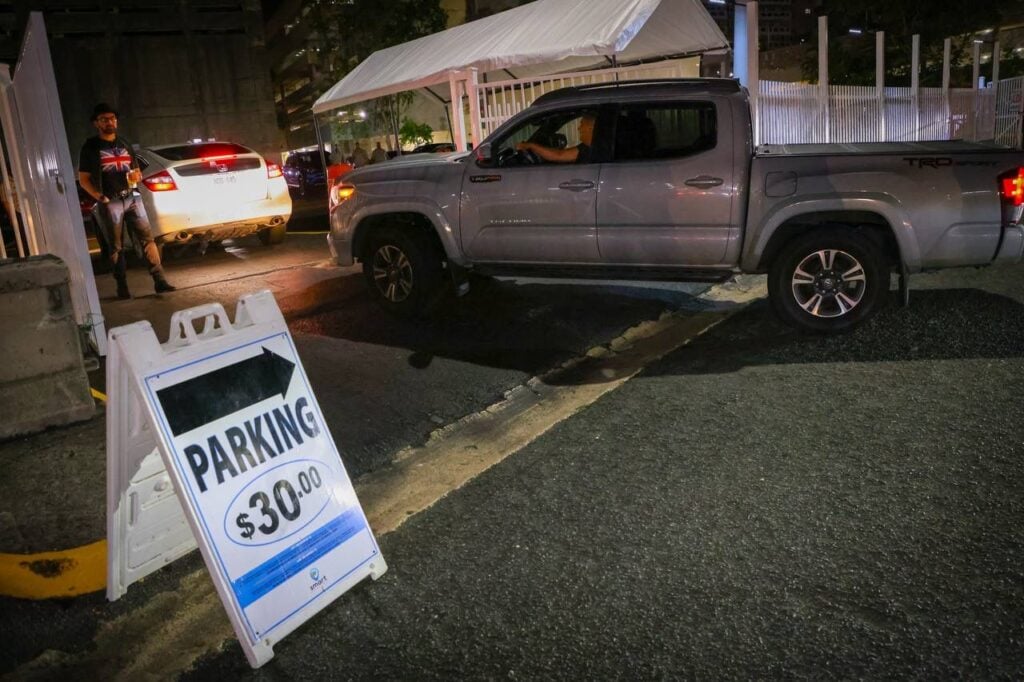Un letrero de estacionamiento indica un costo de $30.00 y señala hacia la izquierda. En la imagen, un hombre con una camiseta negra y un diseño de la bandera británica se encuentra junto a un vehículo blanco que está saliendo del área de estacionamiento. A la derecha, se observa una camioneta gris estacionada, mientras que el entorno es nocturno, con luces de la ciudad de fondo.