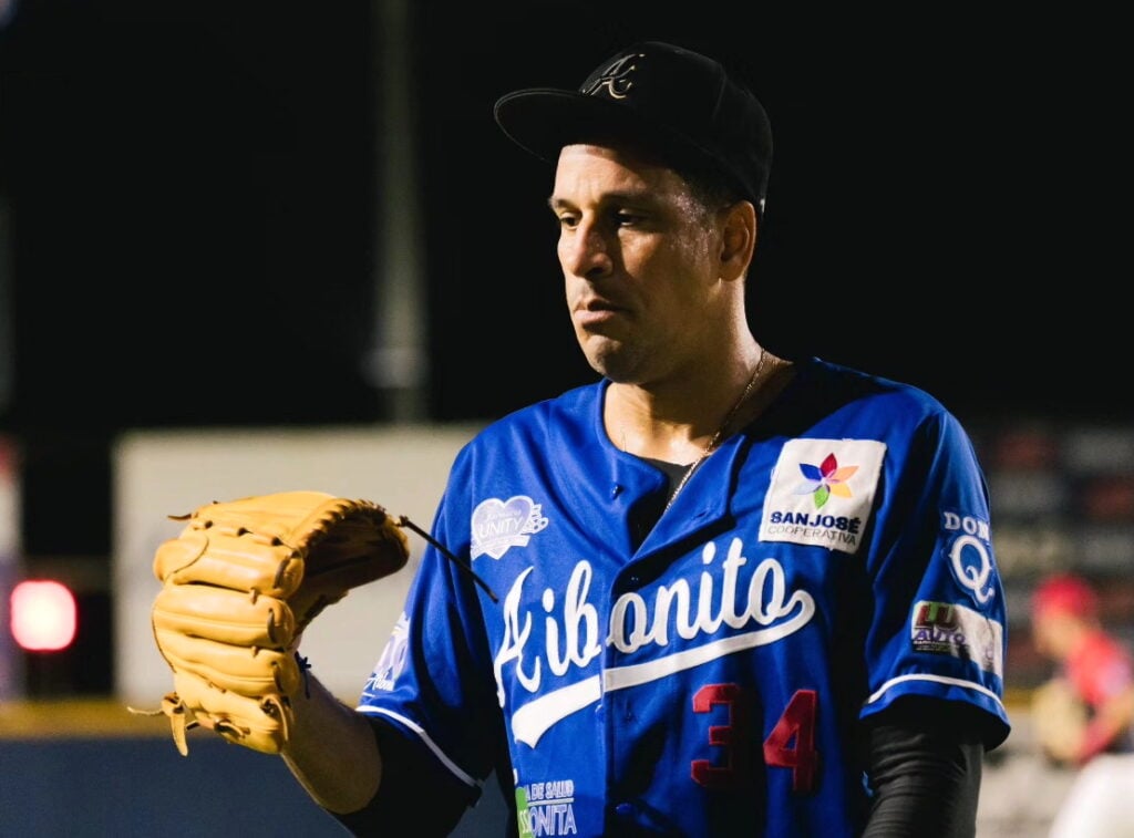 Un jugador de béisbol viste una camiseta azul con el nombre "Cibao" y el número 34. Sostiene un guante de béisbol en su mano izquierda y parece estar concentrado, con una expresión seria. La imagen se toma en un campo de béisbol durante la noche, con luces de fondo que iluminan el entorno.